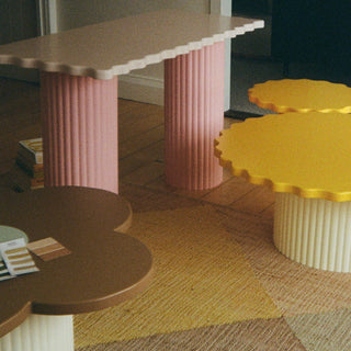 A styled interior scene showing a set of modern, sculptural nesting tables. The collection includes a cloud-shaped coffee table in the foreground with a dark brown top and white ribbed base, a rectangular table in the background with pink ribbed columns and a scalloped light top, and two overlapping round tables on the right with bright yellow scalloped tops and white ribbed bases.