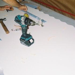 A teal and black cordless drill with a bit attached, resting on a white surface with a hammer nearby. A person's hands are visible holding tape near the top of the frame, and a brown tent-like fabric is in the background.