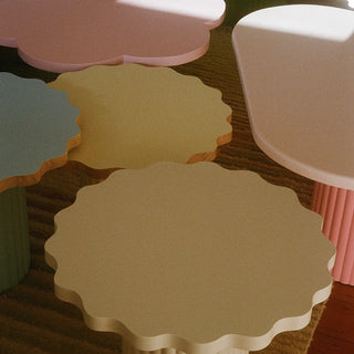 A set of pastel-colored, scalloped-edge nesting tables or stools arranged together on a textured brown rug. The visible pieces include a large cream/beige table in the foreground, a pale yellow one behind it, a pink cloud-shaped piece at the top, a light blue piece on the left, and a white ribbed stool on the right.