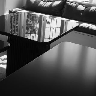 A modern black coffee table with a glossy, reflective glass top and a ribbed black pedestal base, shown in a living room setting with a dark sofa in the background.