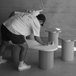 A person in a white t-shirt and dark shorts leans over a ribbed white table in a minimalist studio setting with matching ribbed pedestal tables.