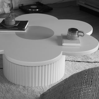 A white flower-shaped coffee table with a ribbed cylindrical base, styled with books and a ceramic mug in a home interior setting.