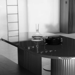 A modern glossy black oval dining table with fluted pedestal legs, shown in a minimalist interior setting. A clear glass decanter and a stack of black plates rest on the reflective tabletop.