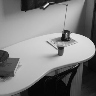 A modern curved white desk in a minimalist room setting, styled with a silver adjustable desk lamp, a coffee cup, a book, and a magazine. A black wooden chair is partially visible underneath.