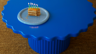 A small slice of layered cake with lit birthday candles sits on a light blue plate, placed on a bright blue scalloped pedestal table over a brown carpeted floor.