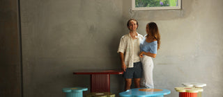 A couple stands behind a set of colorful ribbed modern furniture pieces in a minimalist concrete room. The furniture includes a red rectangular table, several cylindrical stools in light blue, olive green, and red, a light blue oval table, and a dark blue rectangular table with a white top.