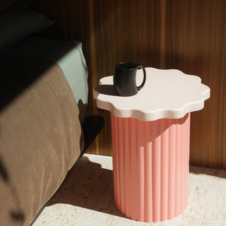 Pink side table with scalloped edge holding a black mug next to a bed.