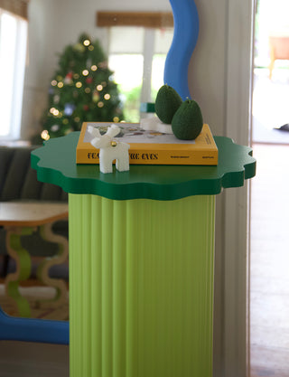 Green decorative stand with a small white figurine and greenery, set against a blurred indoor background.