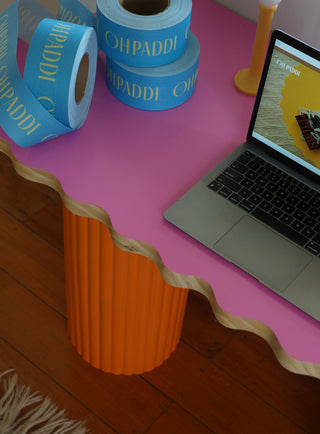 Pink desk with orange legs, laptop, and rolls of blue paper with 'CHI PADDI' branding.